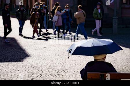 Frankfurt, Deutschland. 22 2021. April: Eine Frau sitzt als Sonnenschirm auf einer Bank auf dem Römerberg unter ihrem offenen Schirm, während eine Gruppe junger Menschen vor ihr entlang gehen, von denen einige keinen Mund-/Nasenschutz tragen, obwohl dies hier obligatorisch ist. Foto: Frank Rumpenhorst/dpa Quelle: dpa picture Alliance/Alamy Live News Stockfoto