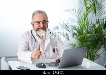 Leitender bärtiger Arzt, der lächelnd mit Daumen nach oben in seinem Arzt arbeitet Büro Stockfoto