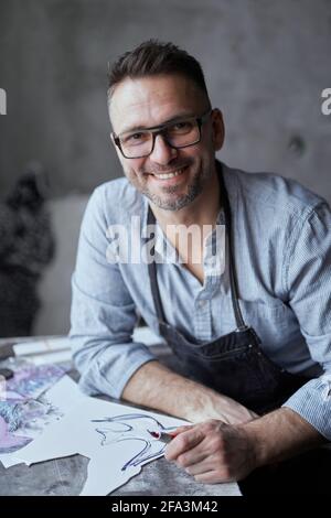 Bärtiger Handwerker mittleren Alters in einer Brille, die auf einem Hintergrund des Arbeitsplatzes sitzt. Linolschnitt-Meister in Schürze lächelnd und gerade in die Kamera schauend und lächelnd. Hochwertige Fotos Stockfoto