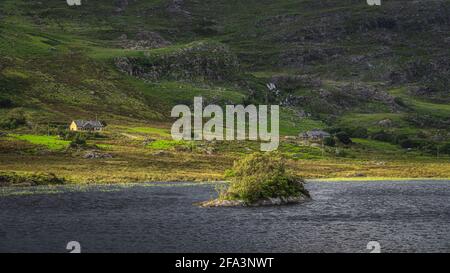 Kleine felsige Insel mit verdrehten Bäumen am Lough Gummeenduff mit Häusern auf dem Hügel von MacGillycuddys Reeks Mountains, Ring of Kerry, Irland Stockfoto