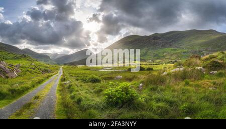 Gerade Landstraße, die durch das Black Valley führt, dramatischer Himmel. Landschaft mit See, Fluss und MacGillycuddys Reeks Mountains, Ring of Kerry, Irland Stockfoto