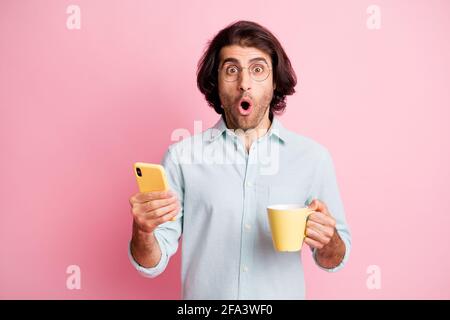Foto von gut aussehenden beeindruckt braunen Haar Kerl halten Tasse Telefon Tragen Sie Brillen blaues Hemd isoliert auf pastellrosa Hintergrund Stockfoto