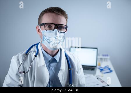 Deutscher europäischer junger Arzt, der in der Klinik am Arbeitsplatz mit müdem Blick auf die Kamera mit Brille und Maske schaut. Thema ist die harte Arbeit von Stockfoto