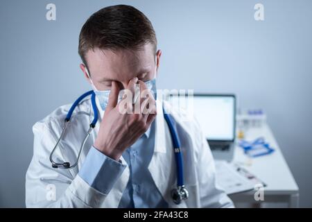 Deutscher europäischer junger Arzt, der in der Klinik am Arbeitsplatz mit müdem Blick auf die Kamera mit Brille und Maske schaut. Thema ist die harte Arbeit von Stockfoto