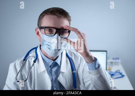 Erschöpfter junger Arzt im Laborkittel zieht Maske und Brille ab, reibt die Augen, hat Kopfschmerzen durch Ermüdung und Überanstrengung durch die Arbeit in schwerer Schicht Stockfoto