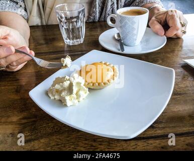 Frauenhand hält Gabel mit Biss von Lemon Tart und Gerinnte Cremekaffee auf dem Tisch neben dem Teller Stockfoto