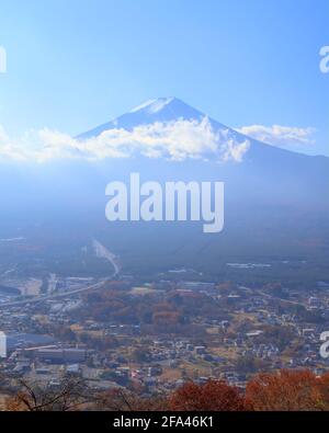 Tagesansicht des Fuji aus der Ferne, teilweise bedeckt von niedrigen Wolken unter einem hellblauen Himmel und mit einem Teil der Stadt Fujiyoshida sichtbar Stockfoto