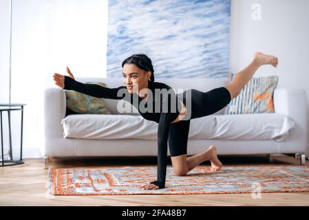 Nettes sportliches afroamerikanisches Mädchen in schwarzer Sportkleidung, Stretching auf dem Boden zu Hause, Fitness, Arme und Beine hochheben, einen gesunden Lebensstil führen, lächeln Stockfoto