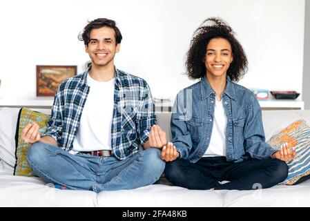 Verliebte Mischling verbringen Zeit miteinander, entspannen und meditieren, während sie in Lotusposition auf dem Sofa sitzen, Stress lindern, die Kamera betrachten und lächeln. Guy und das Mädchen kümmern sich um ihre Gesundheit Stockfoto