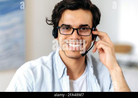 Nahaufnahme eines glücklichen, erfolgreichen jungen hispanischen Mannes mit Brille und Headset, freiberuflicher Mitarbeiter, Callcenter oder Support-Mitarbeiter, der freundlich lächelnd auf die Kamera blickt Stockfoto