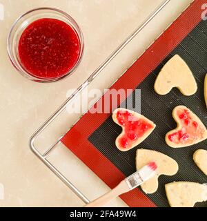 Herzförmige Kekse mit roter Marmelade auf schwarzer Silikonmatte Flach liegend Stockfoto