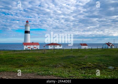 Point Amour Historic Lighthouse am Rande des Atlantischen Ozeans. Der blaue Himmel wird von flauschigen Wolken verdeckt. Ein Gebäude mit zwei Eingängen. Stockfoto