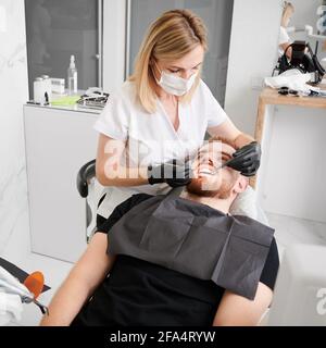 Ansicht von oben auf dem Zahnarztstuhl liegend, mit schwarzem Lätzchen, Zahnärztin, die Zahnarztcheck mit Zahnwerkzeugen durchführt, schwarze Handschuhe, Gesichtsmaske und Uniform trägt. Konzept der Zahnmedizin Stockfoto