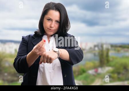 Geschäftsfrau mit ernstem Ausdruck in elegantem, legerem Büroanzug Zeit bedeutet Geldwert Geste zeigt Armbanduhr auf Stadt und Natur-Bac Stockfoto