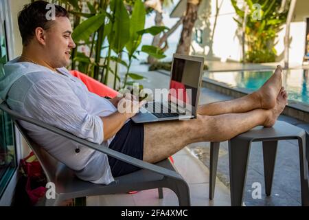 Remote Work Konzept. Mann mit Laptop sitzt am Pool in einem tropischen Land. Stockfoto
