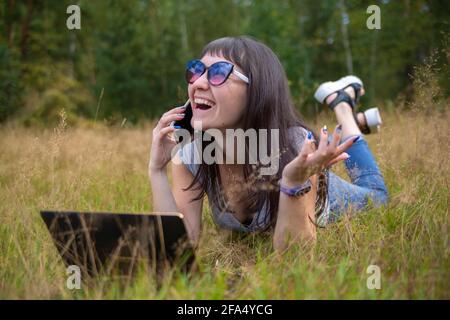 Junge Frau spricht emotional am Telefon auf dem Rasen an einem sonnigen Tag. Glückliche Emotion im Gesicht Stockfoto