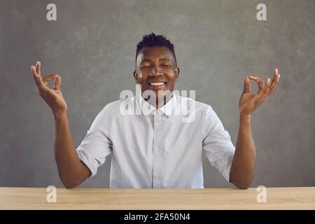 Glücklicher, ruhiger schwarzer Mann, der mit Augen am Schreibtisch sitzt und meditiert Geschlossen und lächelnd Stockfoto