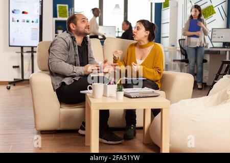 Geschäftskollegen streiten sich, während der Arbeitszeit auf der Couch sitzend, sich gegenseitig in die Wange nehmen, während verschiedene Kollegen im Hintergrund Angst vor der Arbeit haben. Mitarbeiter, die aufgrund von Fehlern aus dem Projekt Konflikte haben Stockfoto