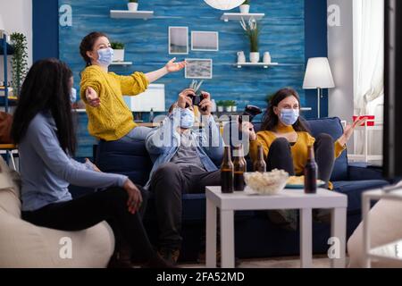 Fröhliche Gruppe multiethnischer Freunde, die den Sieg gegen multiethnische Freunde im Wohnzimmer mit Gesichtsmaske feiern und während des globalen Ausbruchs mit Covid die soziale Distanz bewahren. Neue normale Party. Stockfoto