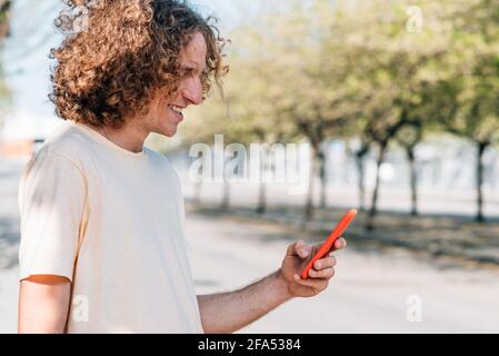 Horizontales Porträt eines rothaarigen jungen Mannes, der ein rotes Smartphone auf der Straße benutzt. Er hat lockiges Haar, lässige Kleidung und lächelt Stockfoto