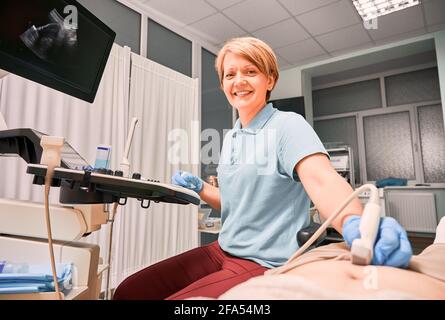 Sonograf mit sterilen Handschuhen, der den Patienten mit einem Ultraschallgerät untersucht. Ärztin, die die Kamera anschaut und lächelt, während sie Ultraschalluntersuchungen im gynäkologischen Kabinett durchführt. Stockfoto