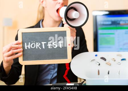 Junge Frau oder Akustiker, die ein Brett oder eine Tafel mit Werbung für einen Hörtest hält, daneben gibt es eine Auswahl an Hörgeräten Stockfoto