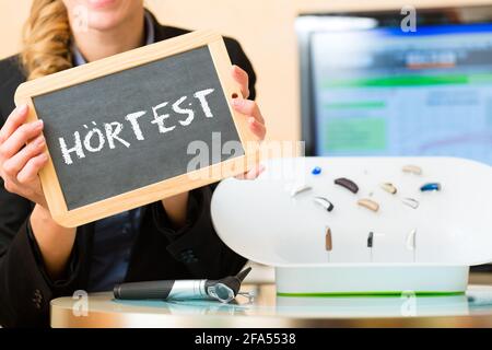 Junge Frau oder Akustiker, die ein Brett oder eine Tafel mit Werbung für einen Hörtest hält, daneben gibt es eine Auswahl an Hörgeräten Stockfoto