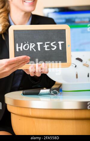 Junge Frau oder Akustiker, die ein Brett oder eine Tafel mit Werbung für einen Hörtest hält, daneben gibt es eine Auswahl an Hörgeräten Stockfoto