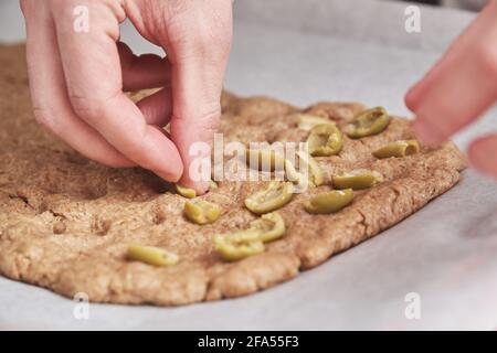 Nicht erkennbarer junger Kaukasier, der Oliven auf einen Teig legt. Er macht Focaccia. Stockfoto