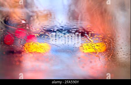 Detail von Regentropfen auf dem Glas eines Autos mit Fahrzeuge vor dem Fahrzeug mit aufgeleuchtter Beleuchtung Stockfoto