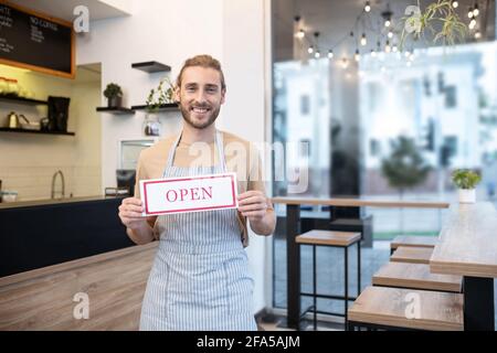 Lächelnder Mann, der ein Schild mit der Aufschrift „Öffnen“ hält Stockfoto