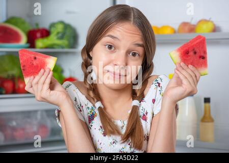 Schöne junge Teenager-Mädchen hält große Wassermelone, während in der Nähe stehen Offener Kühlschrank in der Küche zu Hause Stockfoto