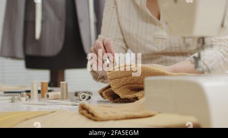 Frau Schneiderin arbeitet in der Werkstatt mit Schneiderpuppe, näht von Hand den Pullover. Nahaufnahme, mit Schneiderpuppe, bunte Kleidung, Stoffe Stockfoto