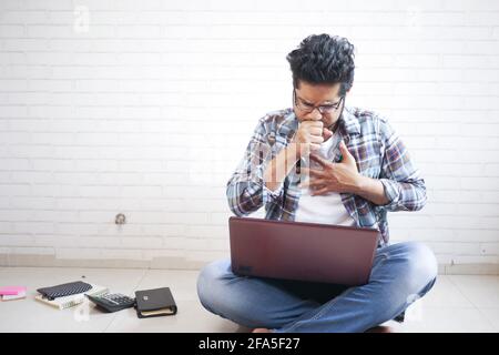 Junger kranker Mann hustet, während er auf einem Laptop sitzt Boden Stockfoto