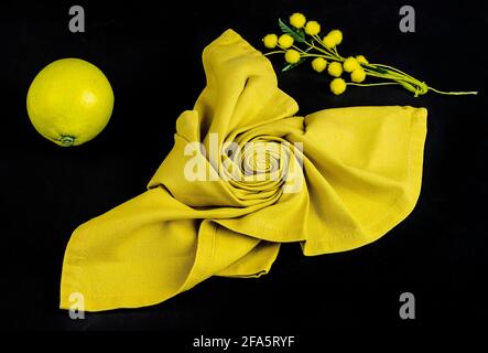 Eine gelbe Stoffserviette mit Blumenform, einer Zitrone und einem Bouquet aus Mimosenblüten auf schwarzem Grund Stockfoto