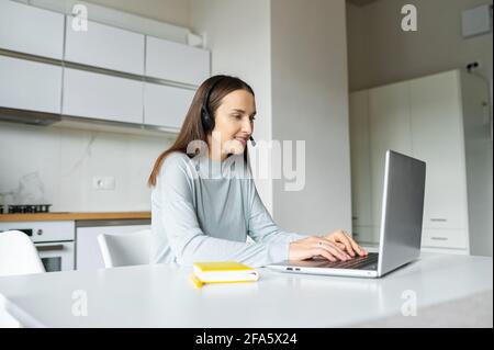 Eine intelligente Studentin, die sich das Online-Webinar ansieht, sitzt eine konzentrierte junge Frau, die ein schnurloses Headset trägt, am Schreibtisch in der Küche zu Hause und nutzt einen Laptop, um online oder an einem anderen Ort zu arbeiten Stockfoto