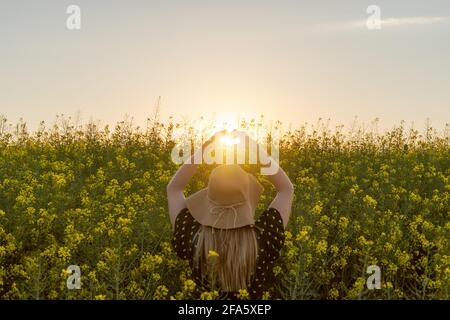 Frau genießen Frühling macht ein Herz mit Händen gegen Sonnenuntergang Sonne Licht in gelben Blumen Feld.Frühling Liebe Konzept, kopieren Raum. Stockfoto