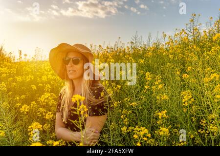 Frau im Stroh Sonnenhut genießen Frühling in gelben Blumen field.Summer Konzept Lifestyle weiblich gegen Sonnenuntergang goldenes Licht in der Natur. Stockfoto