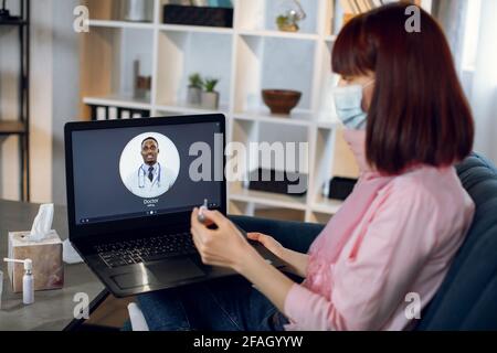 Verschwommenes Bild einer jungen kranken Frau in einer Gesichtsmaske, die auf dem Sofa sitzt und auf den Thermometerbildschirm schaut, der ihre Körpertemperatur überprüft, während sie einen Videoanruf über den pc mit ihrem Arzt führt. Konzentrieren Sie sich auf Notebooks. Stockfoto