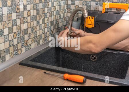 Nahaufnahme der Hände eines Reparaturers in Spezialkleidung, Überprüfung, Reparatur des Wasserhahns in einer modernen Küche, neben einer Kiste mit Werkzeugen. Inklusive Wasserhahn Stockfoto