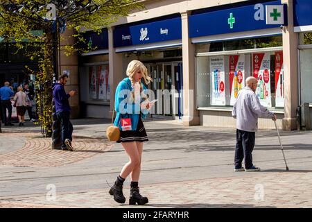 Dundee, Tayside, Schottland, Großbritannien. April 2021. UK Wetter: Warme Frühlingssonne mit einer leichten kühlen Brise durch Nordostschottland mit Temperaturen bis zu 13 Grad Die Sperrung von Covid-19 wird gelockert, da sich die Menschen im Stadtzentrum von Dundee mit den nicht unbedingt notwendigen Geschäften treffen, die am 26. April wieder eröffnet werden sollen. Eine junge modische Frau mit riesigen Plateaustiefeln nutzt die neuen lockeren Sperrbestimmungen und die sonnigen April-Wetternachrichten auf ihrem Smartphone nach einem Einkaufstag. Kredit: Dundee Photographics/Alamy Live Nachrichten Stockfoto