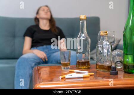 Betrunkene dunkelhaarige Frau, die auf der Couch sitzt, konzentriert sich auf leere Flaschen, Zigaretten. Gesellschaftliches Problem des Alkoholismus Stockfoto