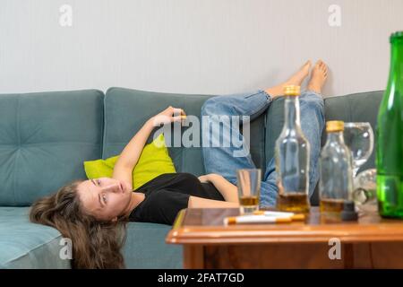 Betrunkene junge dunkelhaarige Frau liegt nach einer Party auf der Couch und schaut an die Decke, leere Flaschen stehen auf dem Tisch Stockfoto