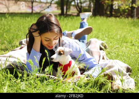 Schöne junge Frau in Casual Outfit auf Picknick im Park mit ihrem entzückenden Jack russell Terrier Welpen, grünes Gras & Laub Hintergrund. Weibliche Besitzerin Stockfoto