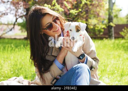 Porträt von attraktiven jungen Frau umarmt niedlichen Jack russell Terrier Welpen im Park, grünen Rasen, Laub Hintergrund. Hipster weiblich in Sonnenbrille Smili Stockfoto