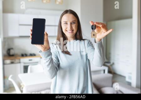 Selektiver Fokus auf ein Smartphone mit leerem Bildschirm und Tasten mit Schlüsselring in Hausform Frau hält. Happy Woman zeigt neue App für die Vermietung von Immobilien, Online-Plattform für den Kauf und Verkauf von Immobilien Stockfoto