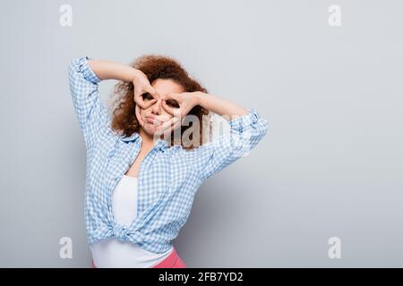 Lockige Frau, die Spaß hat, während sie Ferngläser mit Händen nachahmt Grauer Hintergrund Stockfoto