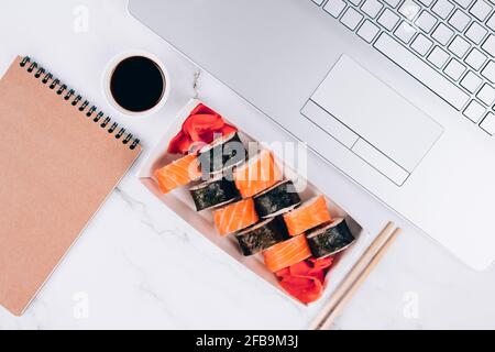 Papierlieferkarton mit Sushi-Rollen mit Lachs auf weißem Marmortisch mit Laptop - Futomaki, Uramaki und Hosomaki. Asiatische Küche Stockfoto