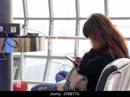 Eine junge Frau an eine Ladestation saßen und auf ihr Smartphone. Aufladen von Handys aus kostenlos Bahnhof im Flughafen. Stockfoto