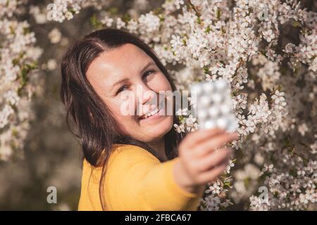 Allergiekonzept, junge Frau mit Pillen oder Medikamenten gegen die starke Allergie in der Hand vor dem Blühen eines Baumes während der Frühjahrssaison, Gesundheitsversorgung Stockfoto